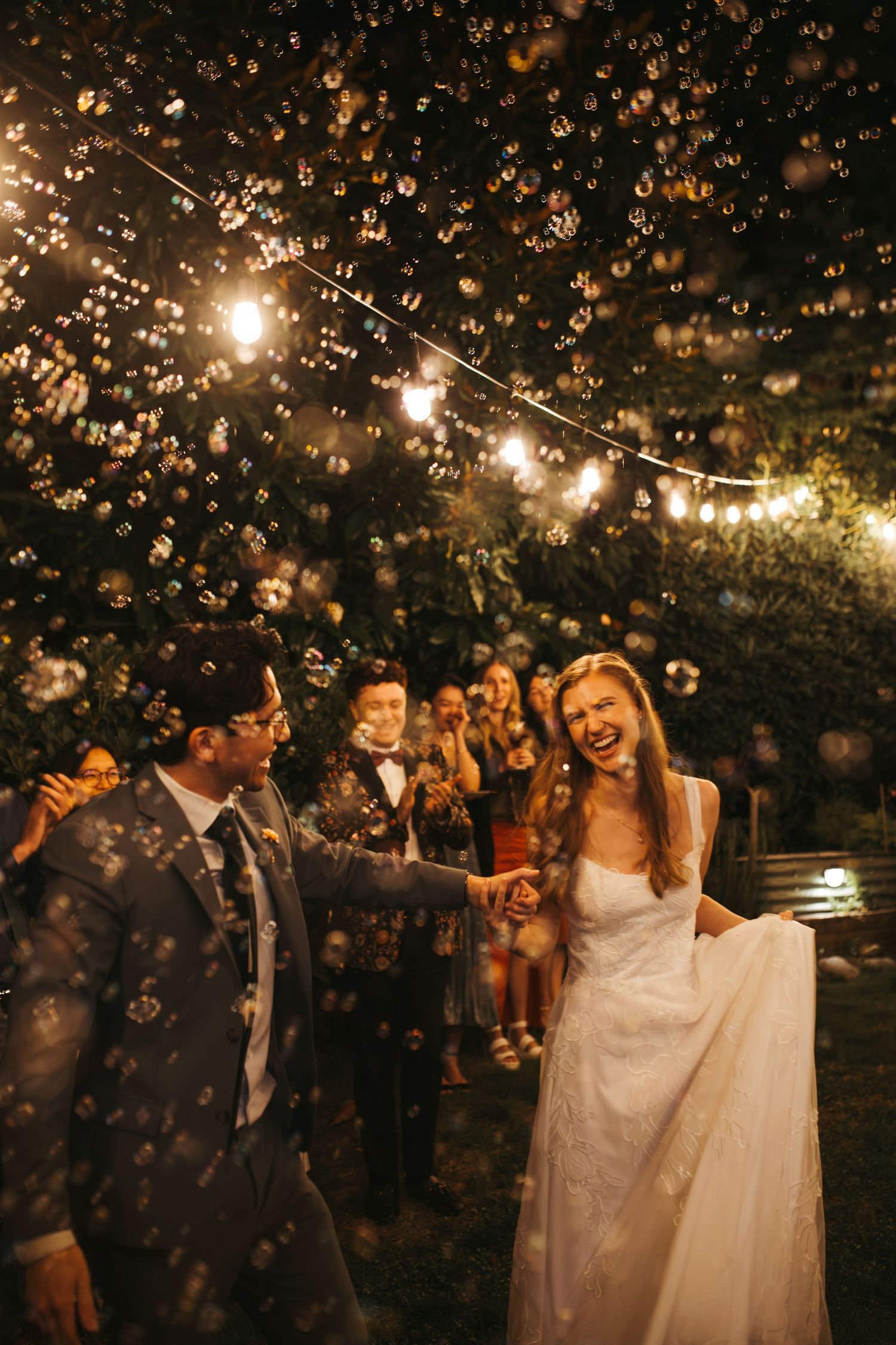 Joyful bride and groom hold hands outdoors at night, surrounded by bubbles and string lights during wedding celebration.