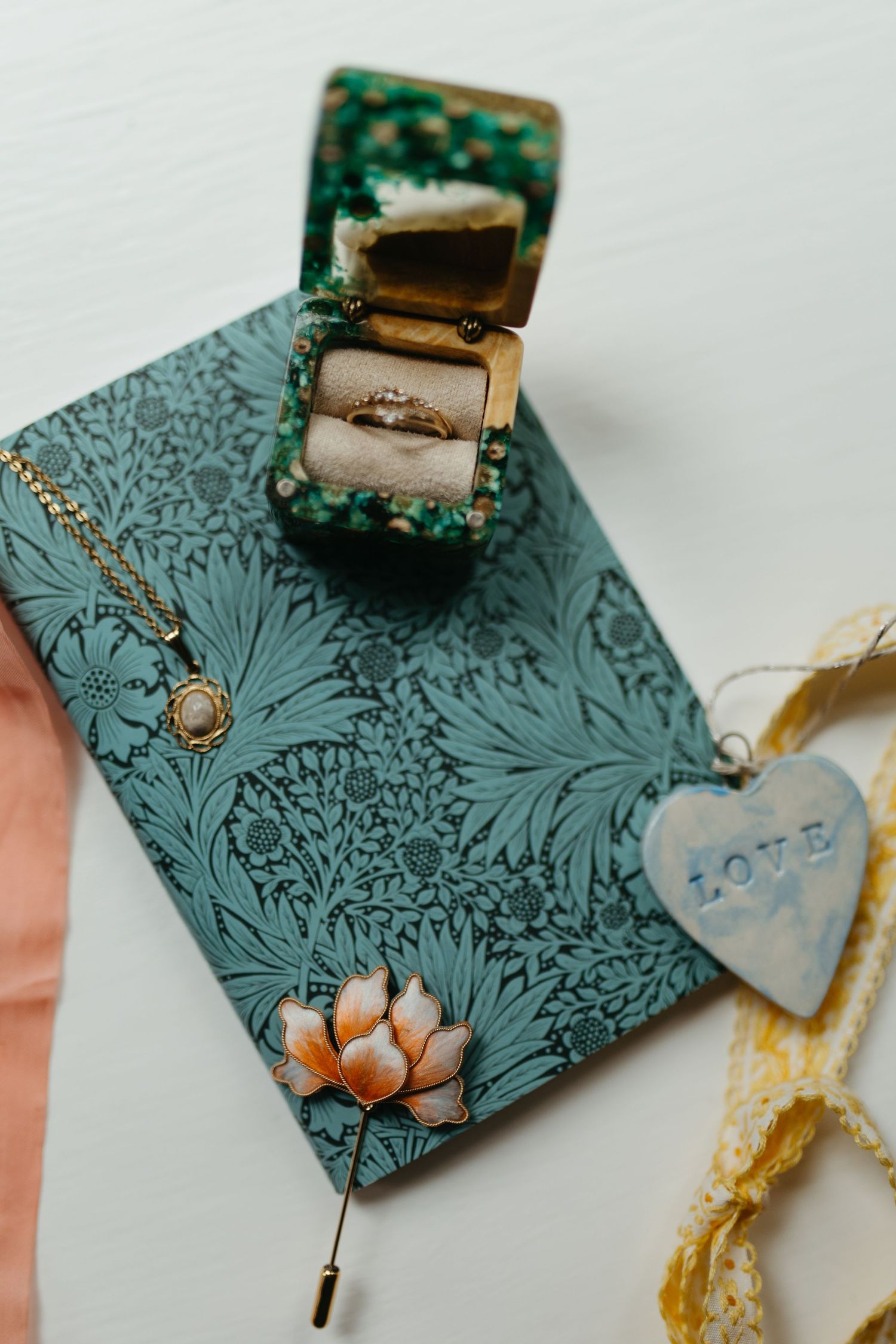Elegant wedding flat lay with teal floral journal, gold ring box, pearl necklace, lotus pin, and heart love tag.