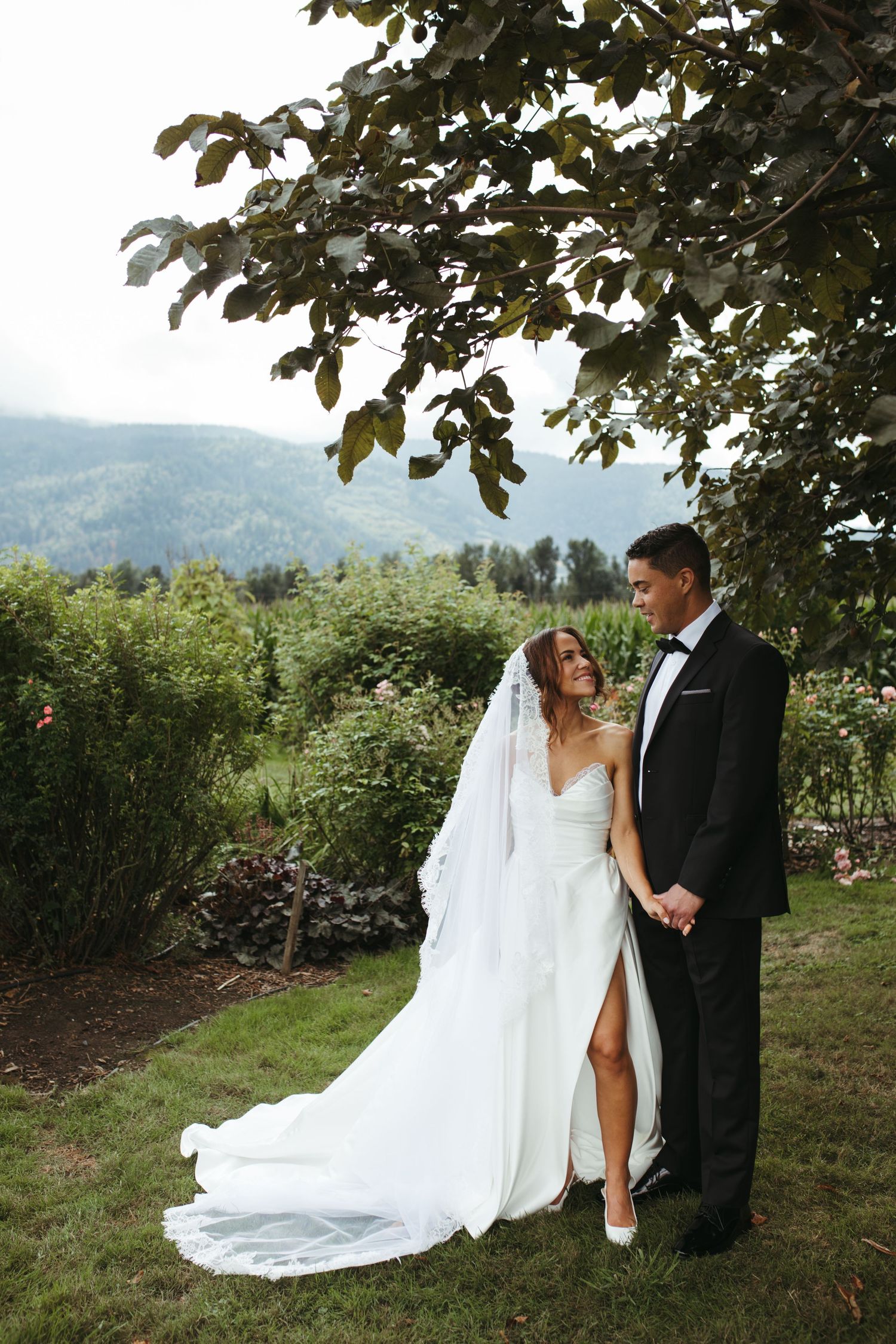 Bride in white gown with veil and groom in black tuxedo stand together outdoors with mountain views.