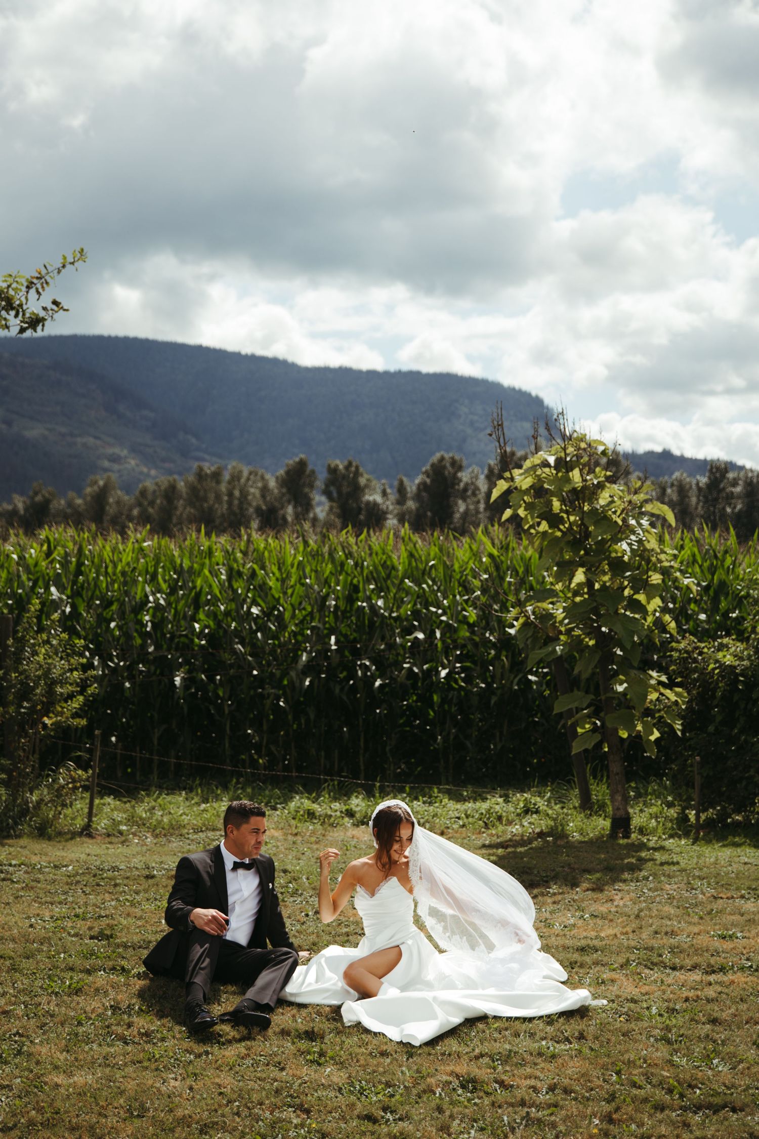 Bride and groom sitting on grass in front of cornfield and mountains on their wedding day.