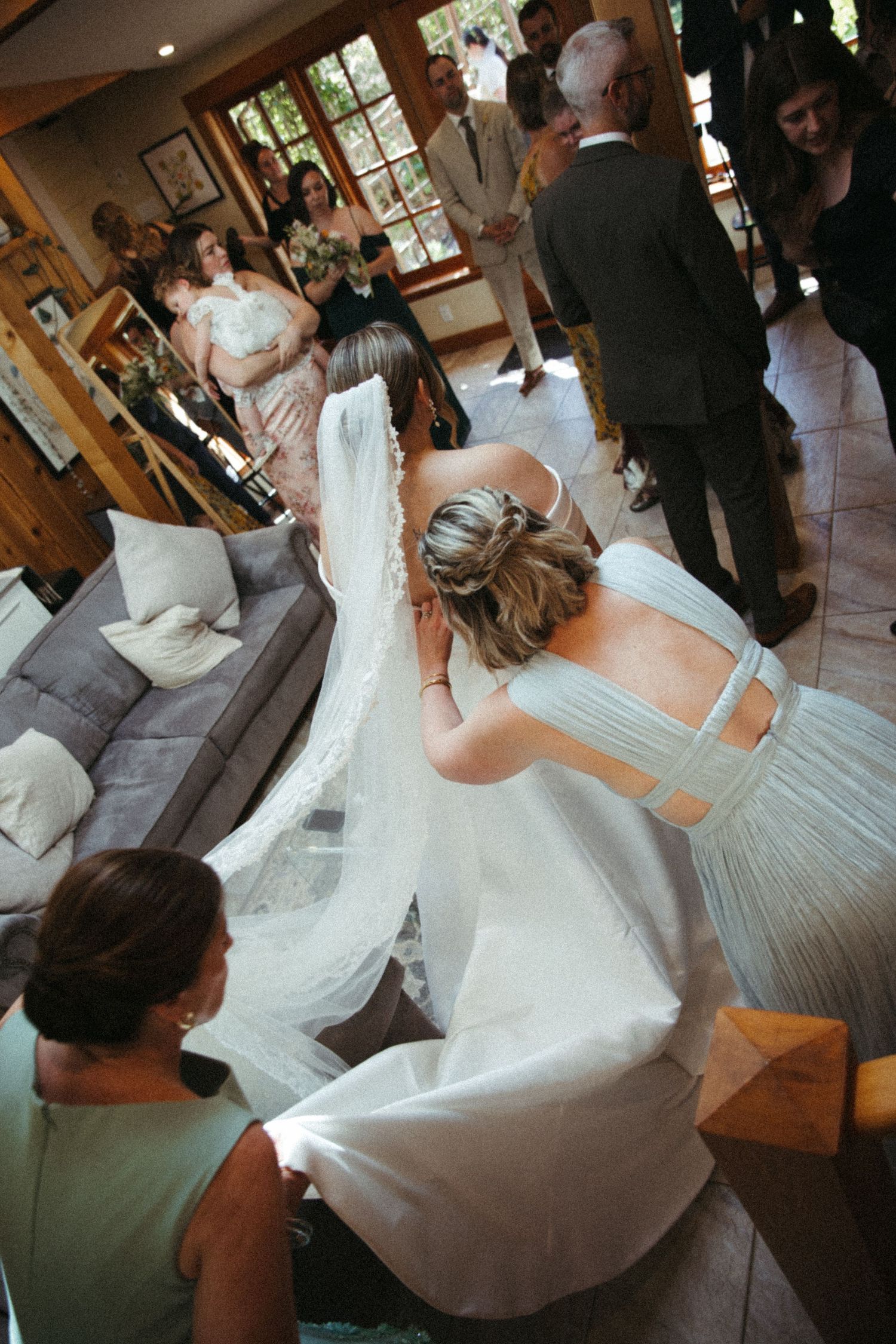 Bridesmaid adjusting bride's veil indoors before wedding ceremony with guests gathered in background.