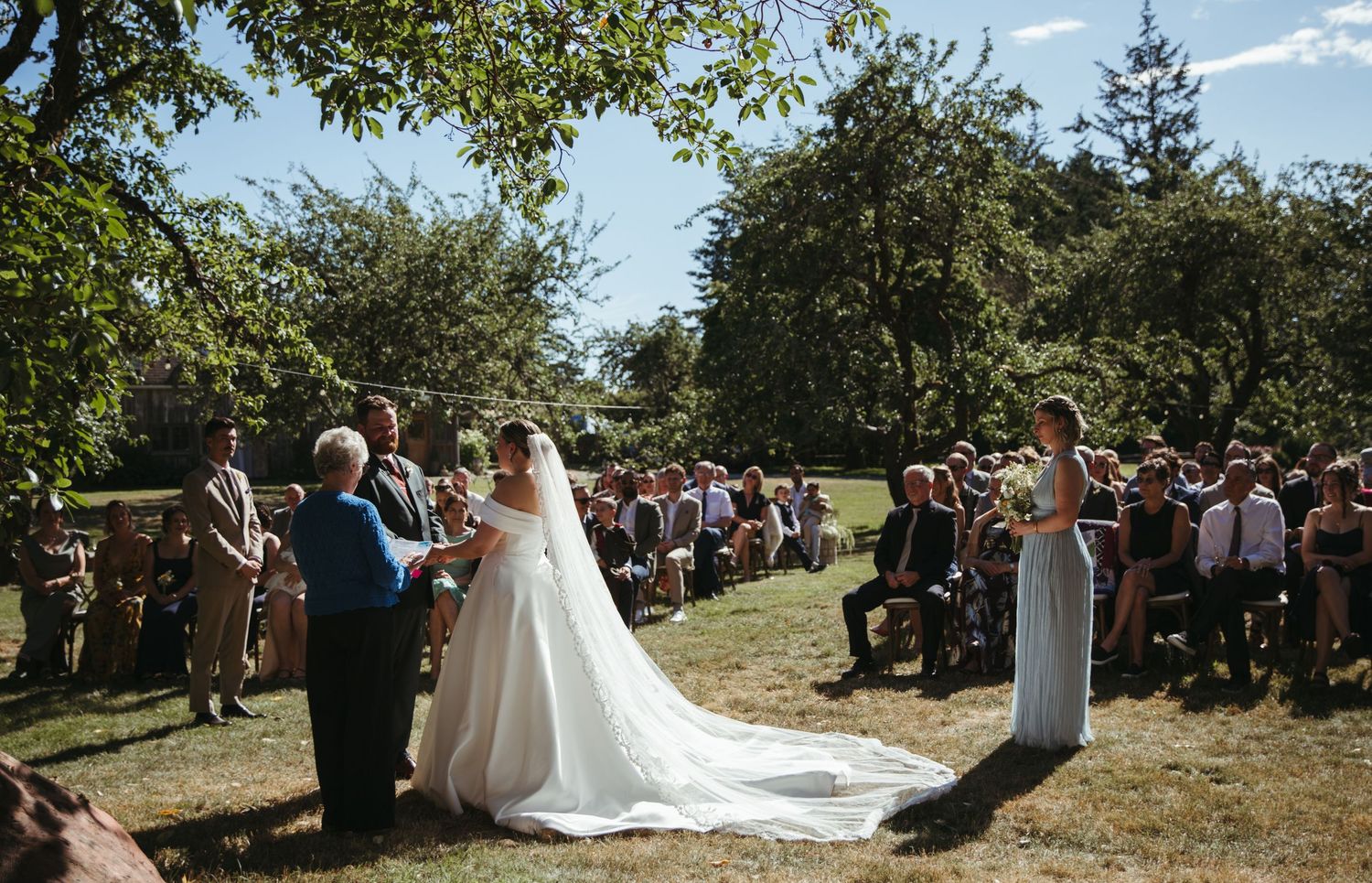 Outdoor wedding ceremony on green lawn with bride in white gown, groom, officiant, and guests seated among lush trees.