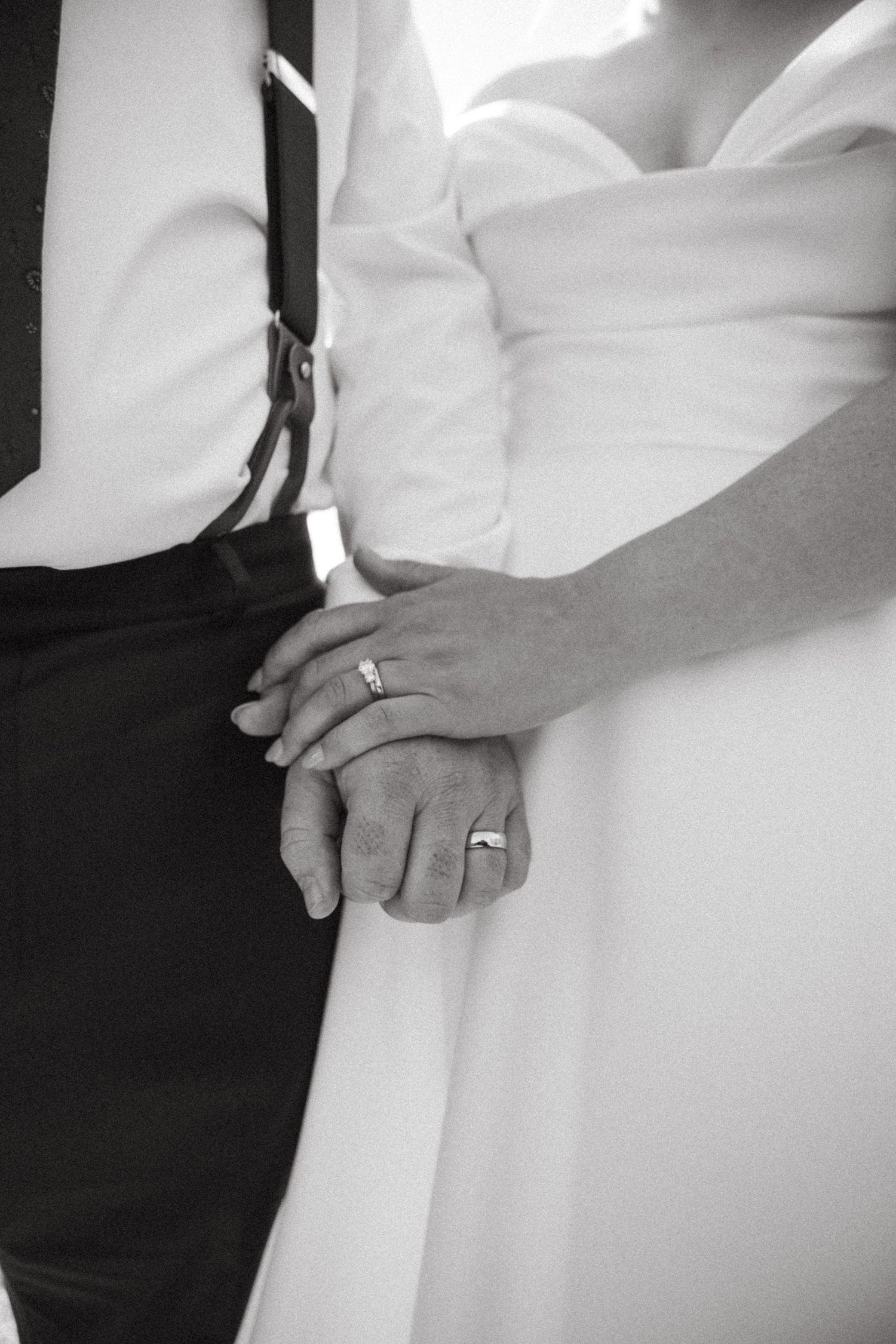 Black and white photo of newlyweds holding hands, showing wedding rings, bride in white gown and groom in suspenders.