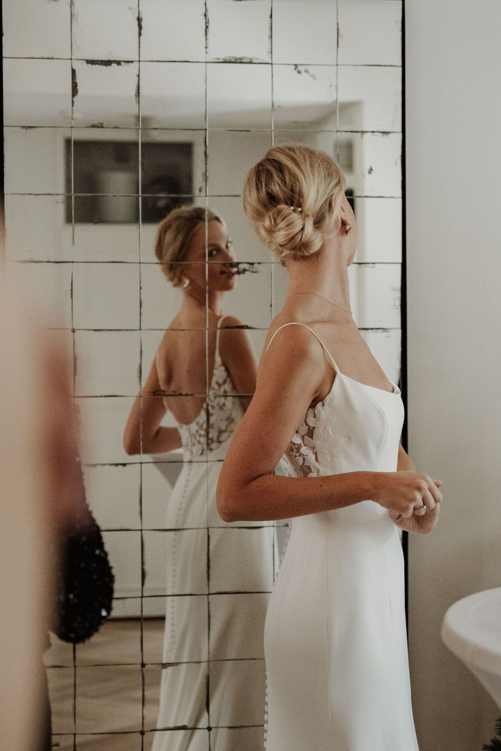 A bride in a sleek white dress looks at her reflection in a tiled bathroom mirror.