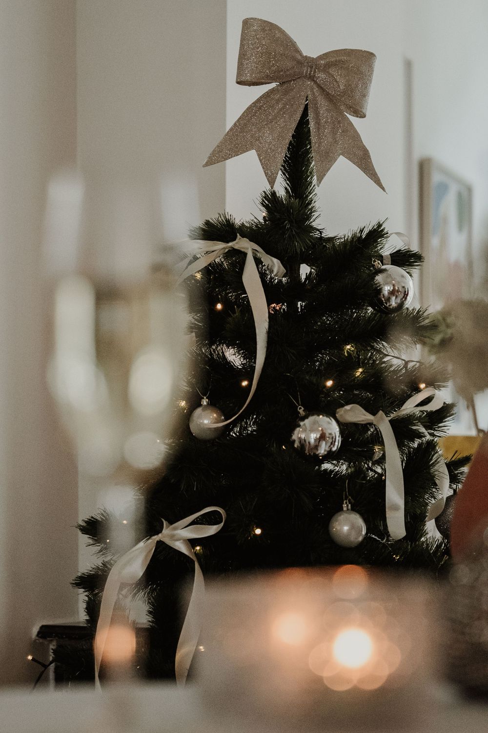 A Christmas tree decorated with silver ornaments and a large burlap bow topper glows with warm bokeh lights in the background.