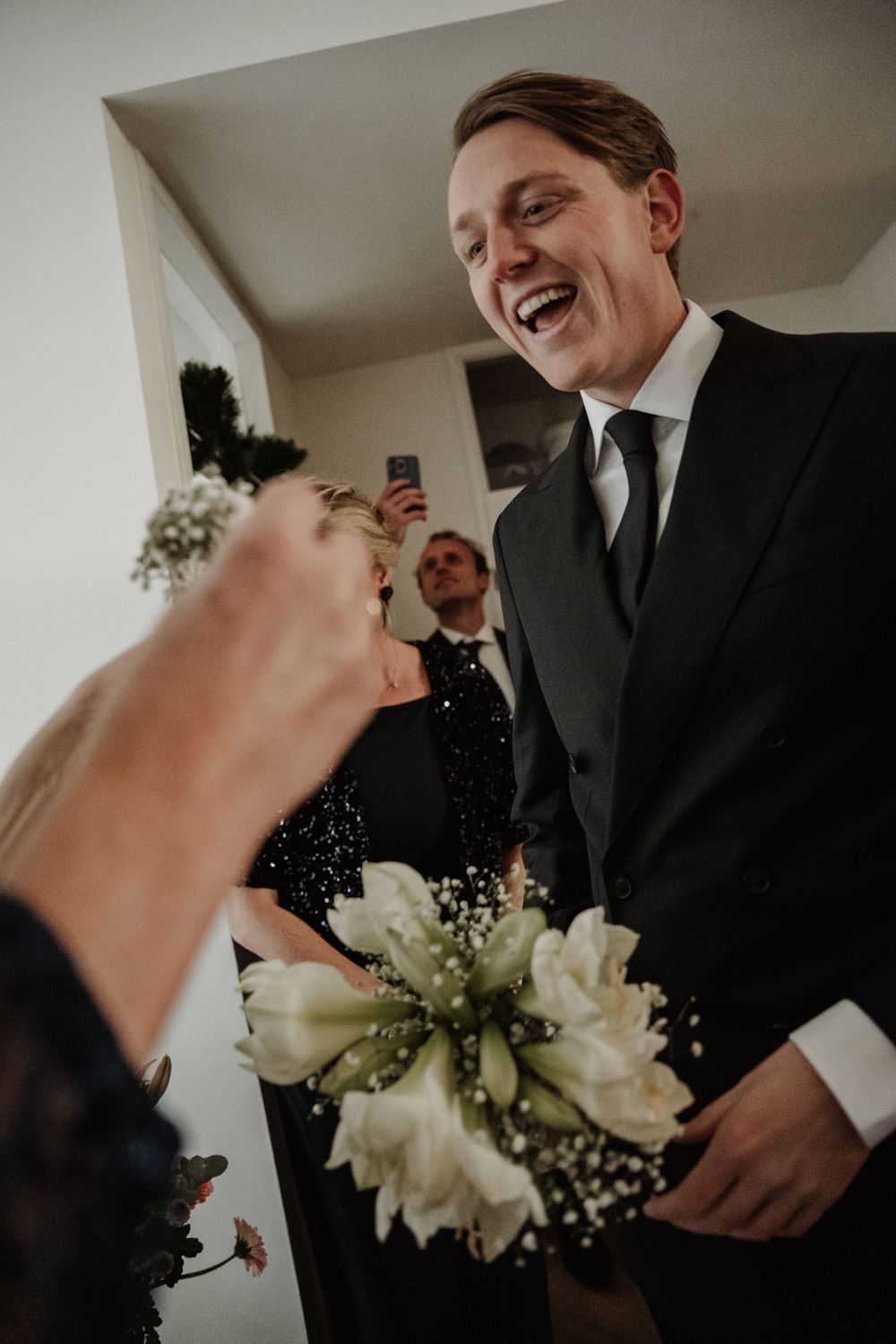 A joyful moment at a wedding with a black suit, tie, and white floral bouquet captured in moody lighting.