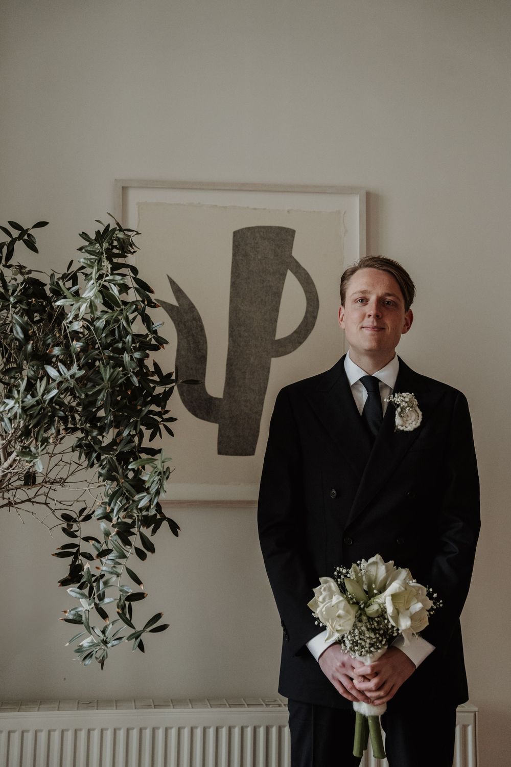 A person in a black suit holds a white bouquet while standing next to a plant and minimalist wall art.