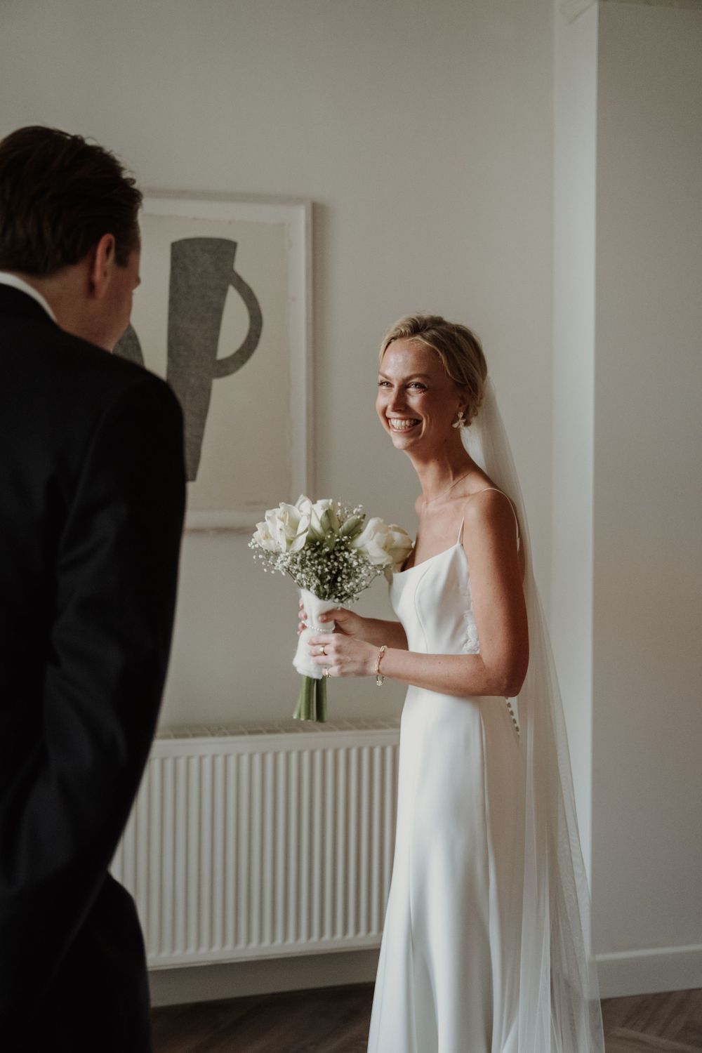A bride in a sleek white gown smiles while holding a bouquet of white flowers during a wedding first look moment.