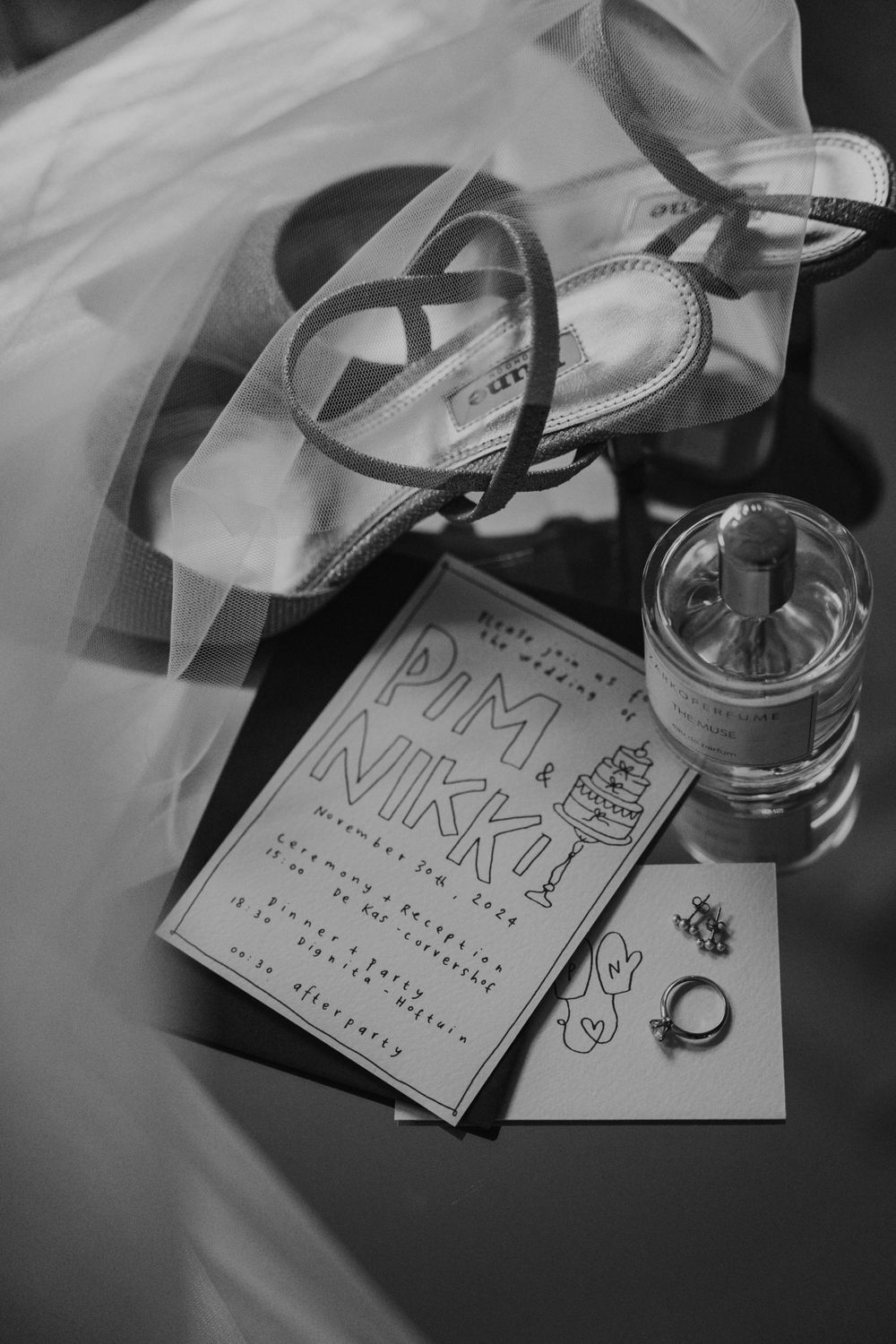 Black and white wedding details including strappy sandals, invitation card, rings and veil with a glass of water.