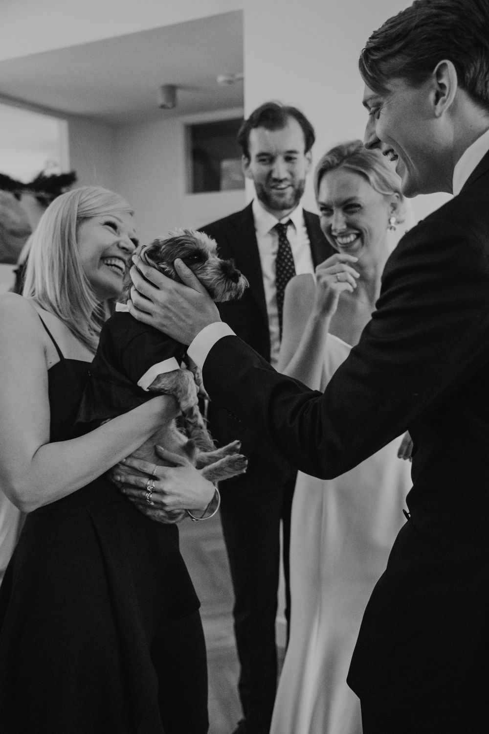 A black and white photo of a group sharing joyful laughter while holding and petting a small dog at an indoor gathering.