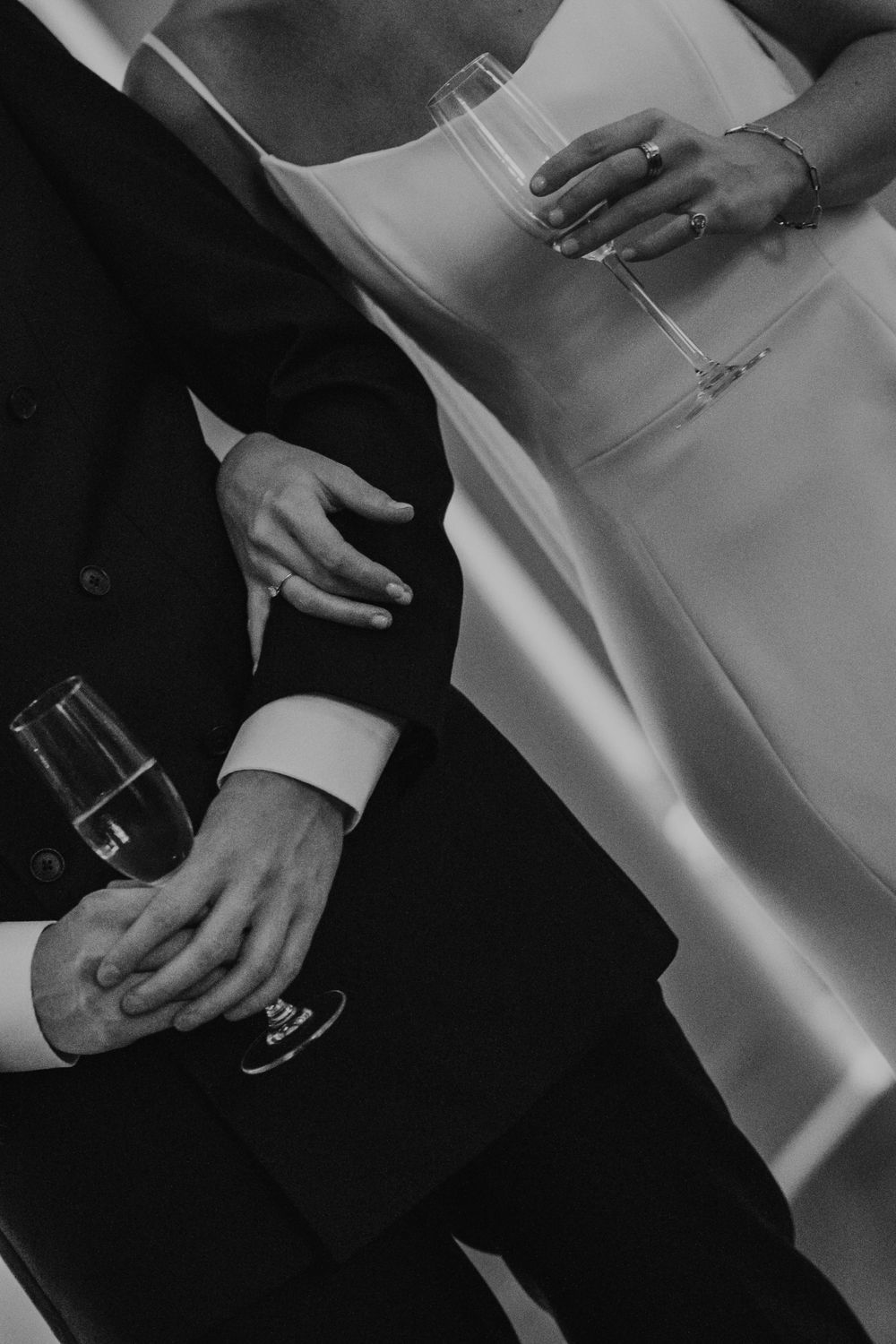 Black and white image of hands holding champagne glasses, contrasting formal black suit against white dress fabric.