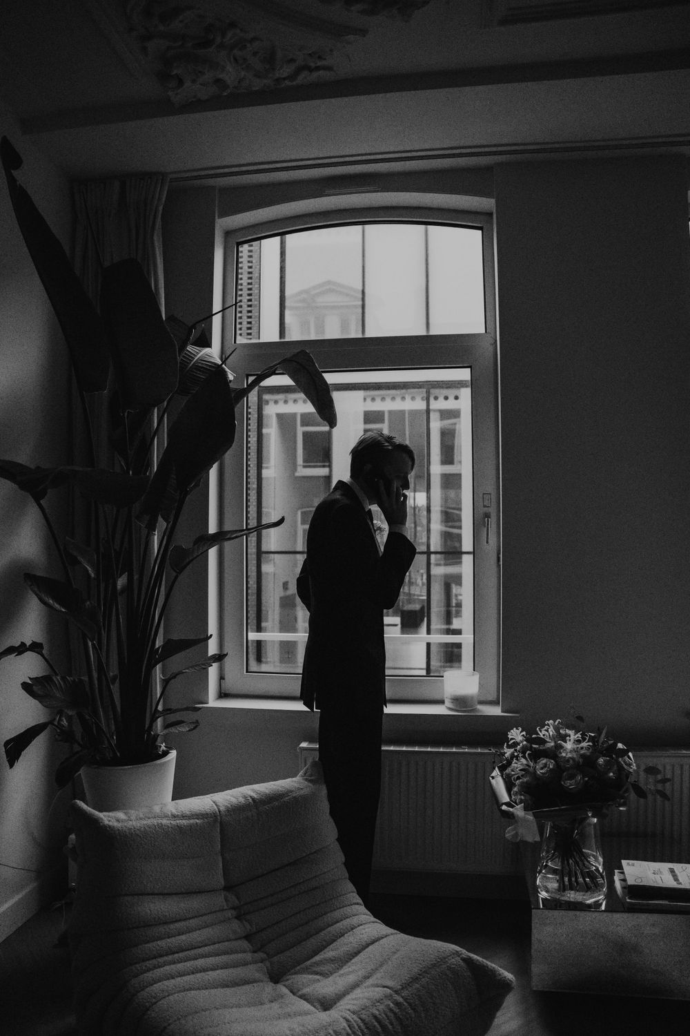 A silhouetted figure stands by a bright arched window in a moody black and white interior with a potted plant and couch.