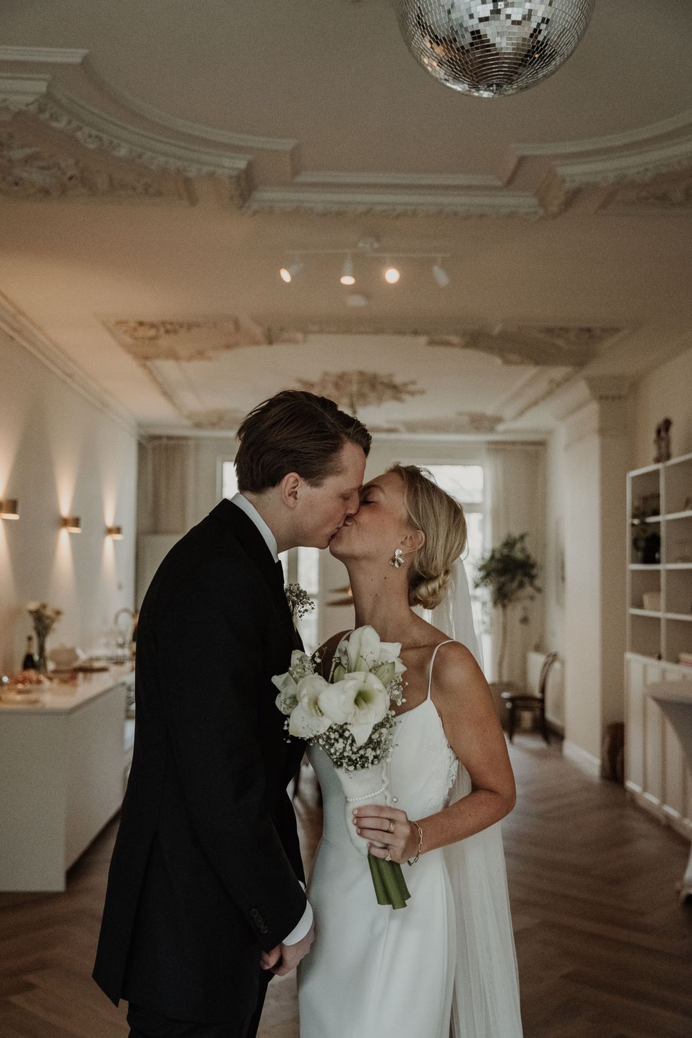 A romantic wedding moment captured in an elegant room with ornate ceiling details and a disco ball overhead.