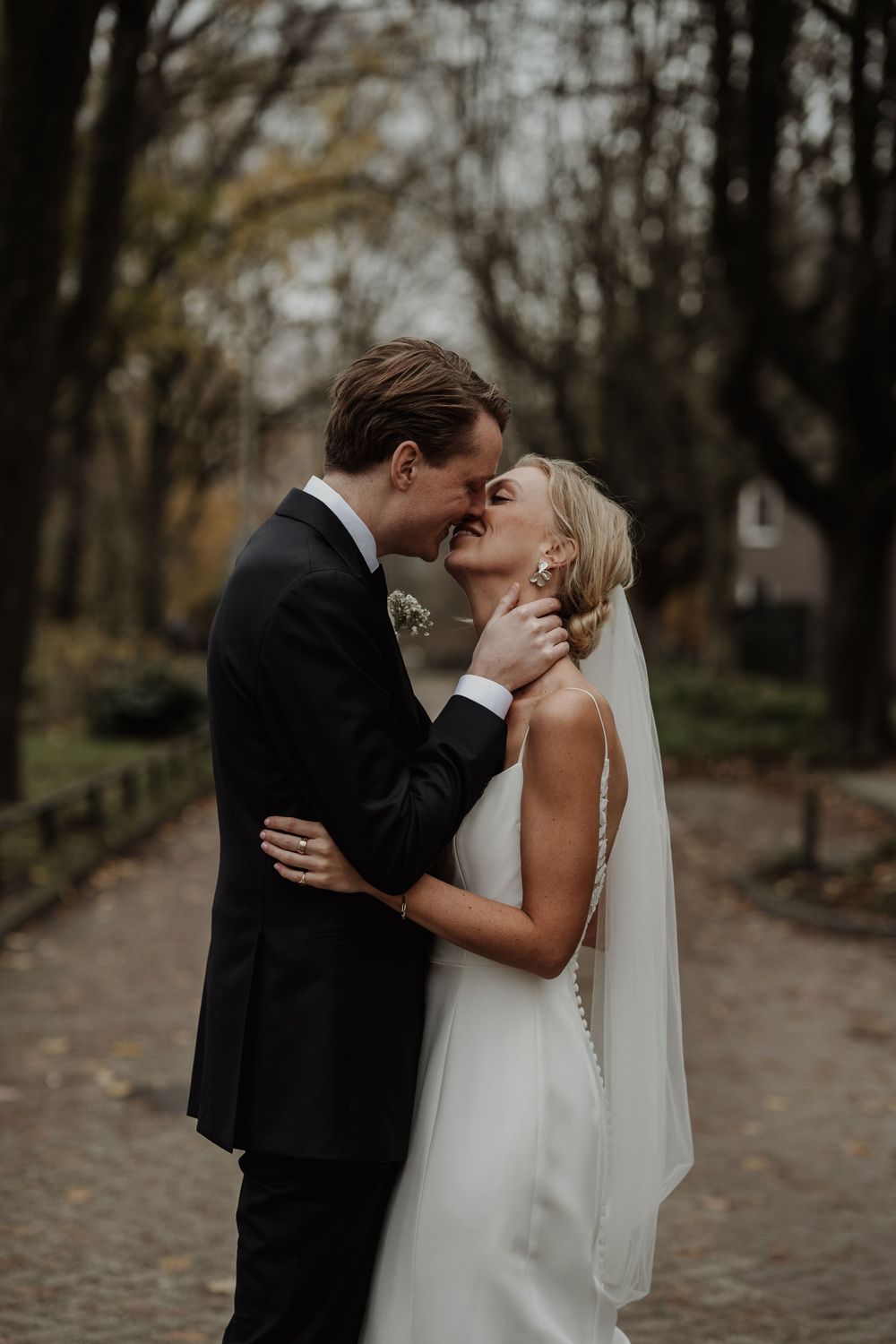 A romantic wedding couple shares a tender kiss on a cobblestone path surrounded by autumn trees.