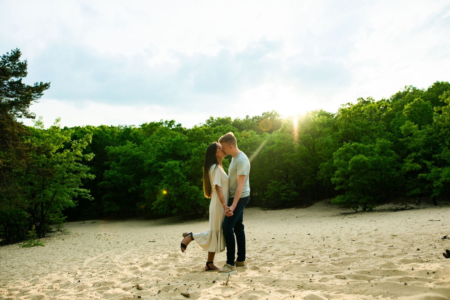 A couple stands together on a dune at sunset with green forest in the background during an engagement shoot.