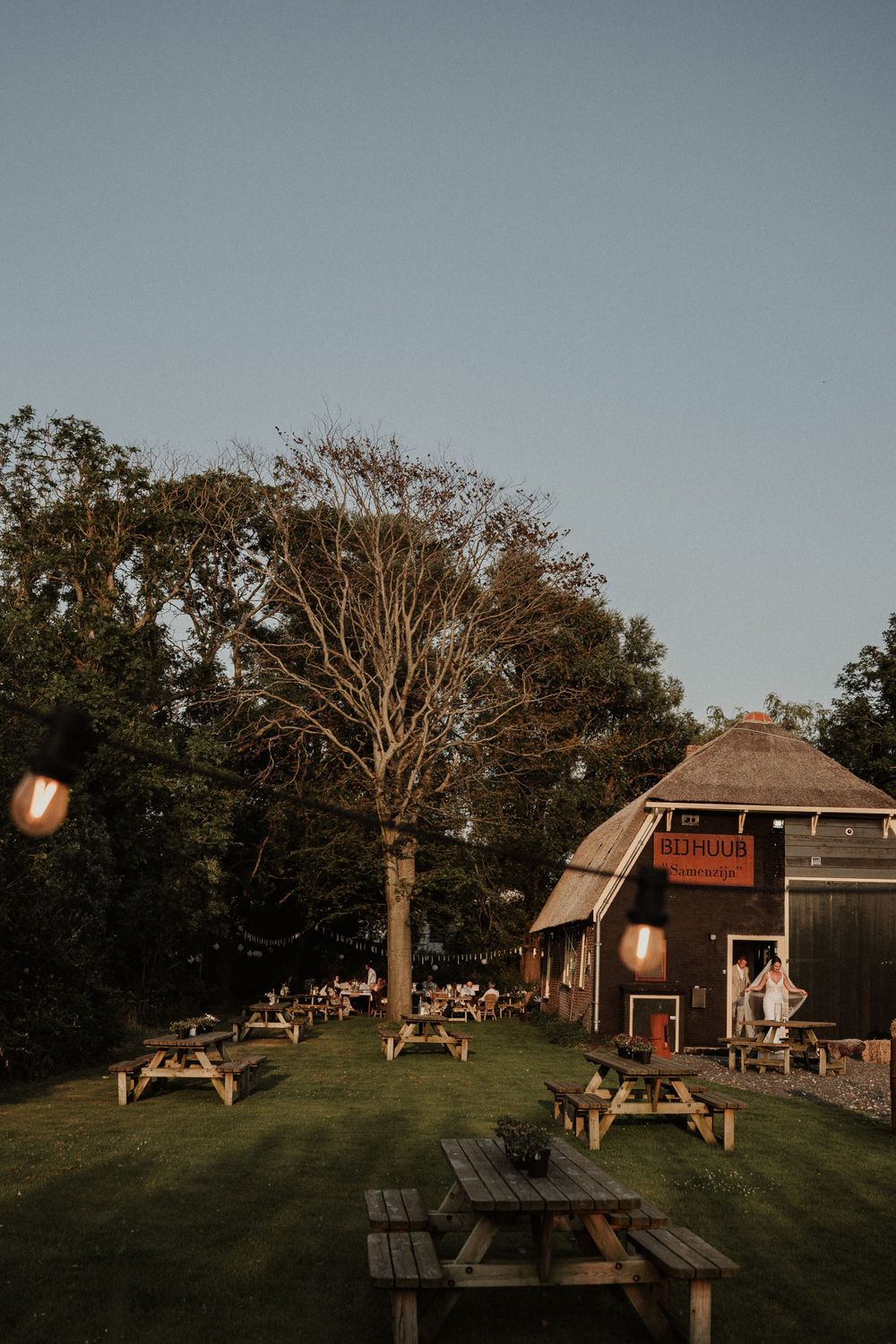 Rustic outdoor beer garden with wooden picnic tables and barn-style building at dusk with warm lighting.
