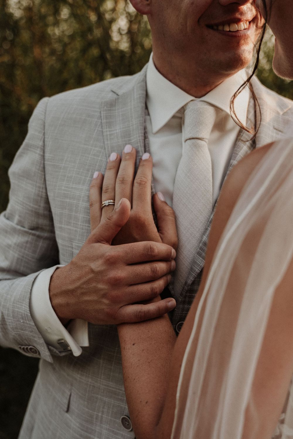 Close-up of newlywed couple's hands showing wedding rings as they embrace, with groom wearing light gray suit and white tie.