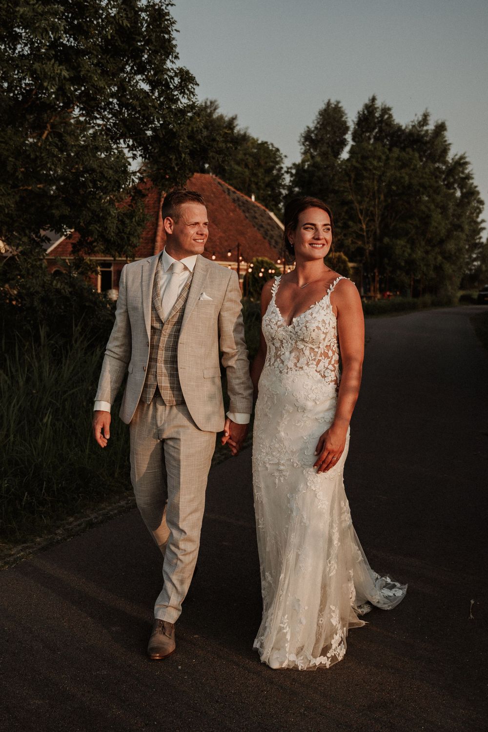 A couple walks together on a country road at sunset, wearing elegant wedding attire.