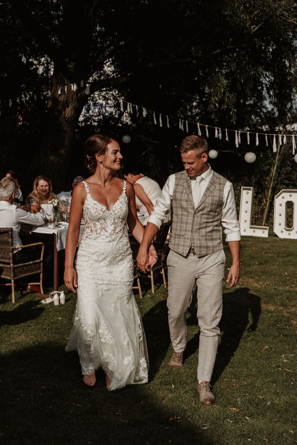 A couple walks hand in hand on their wedding day beneath string lights in a garden setting.