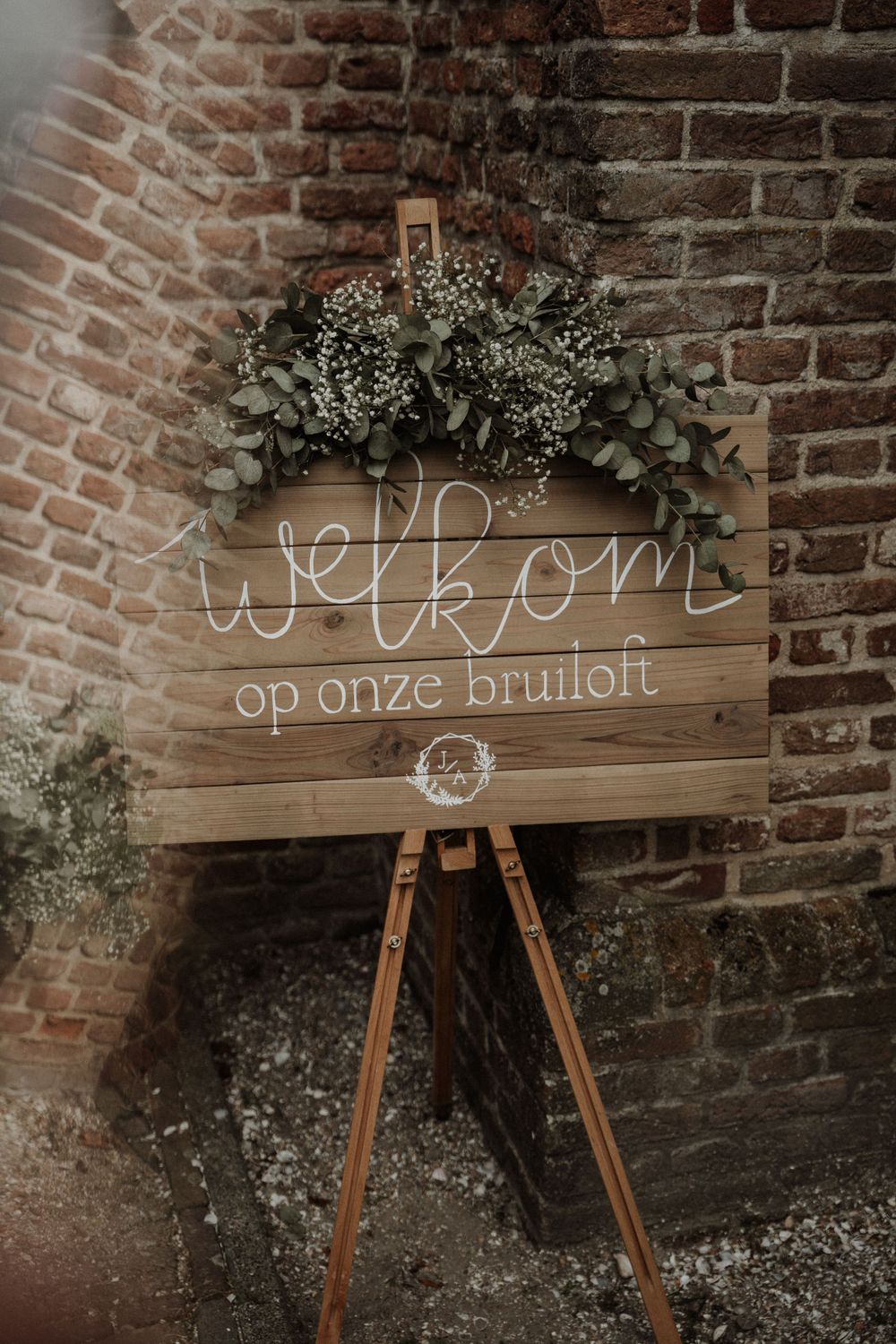 Rustic wooden welcome sign with eucalyptus greenery decoration against brick wall at wedding venue entrance.
