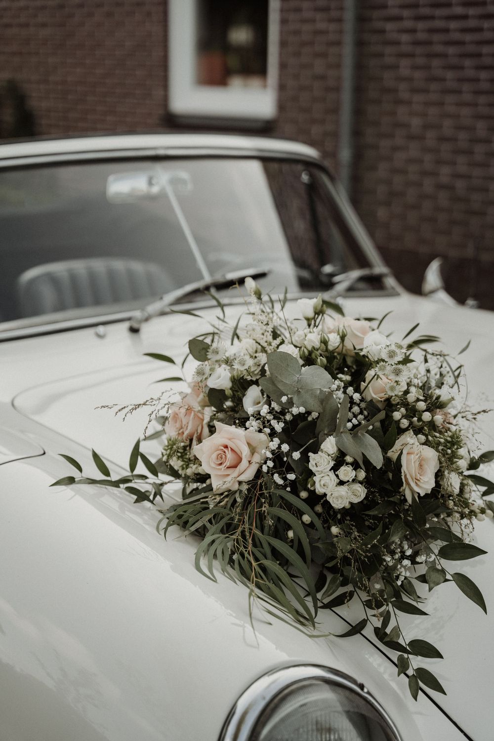 A beautiful floral arrangement with pink roses and baby's breath adorns the hood of a vintage white wedding car.