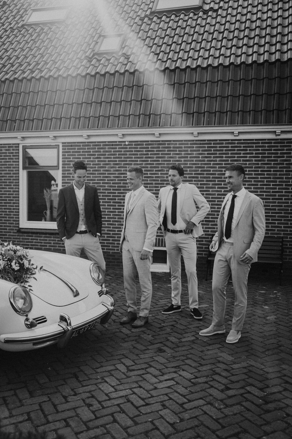 A vintage black and white photo showing a group of men in suits standing next to a classic Porsche on brick pavement.