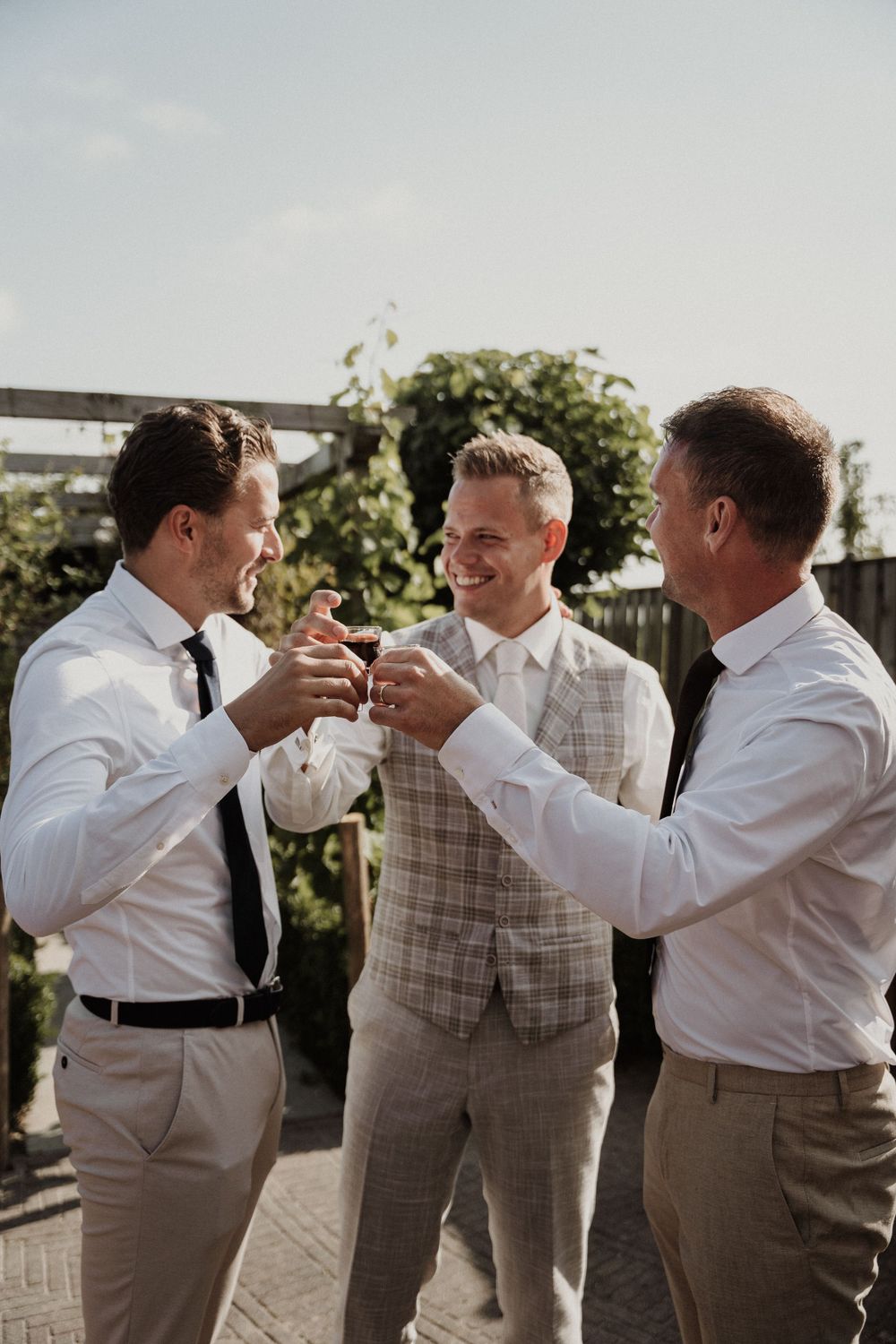 Three groomsmen in formal attire sharing drinks and toasting at an outdoor wedding celebration.