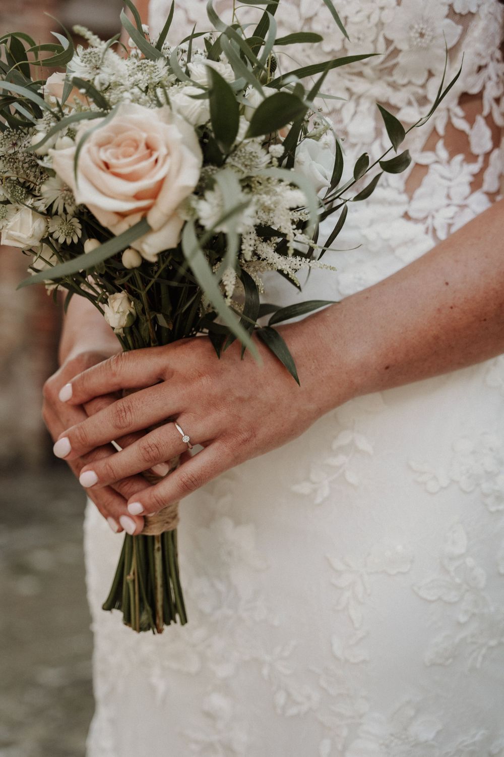 Close-up of hands holding a delicate bridal bouquet with blush roses and greenery against a white lace wedding dress.