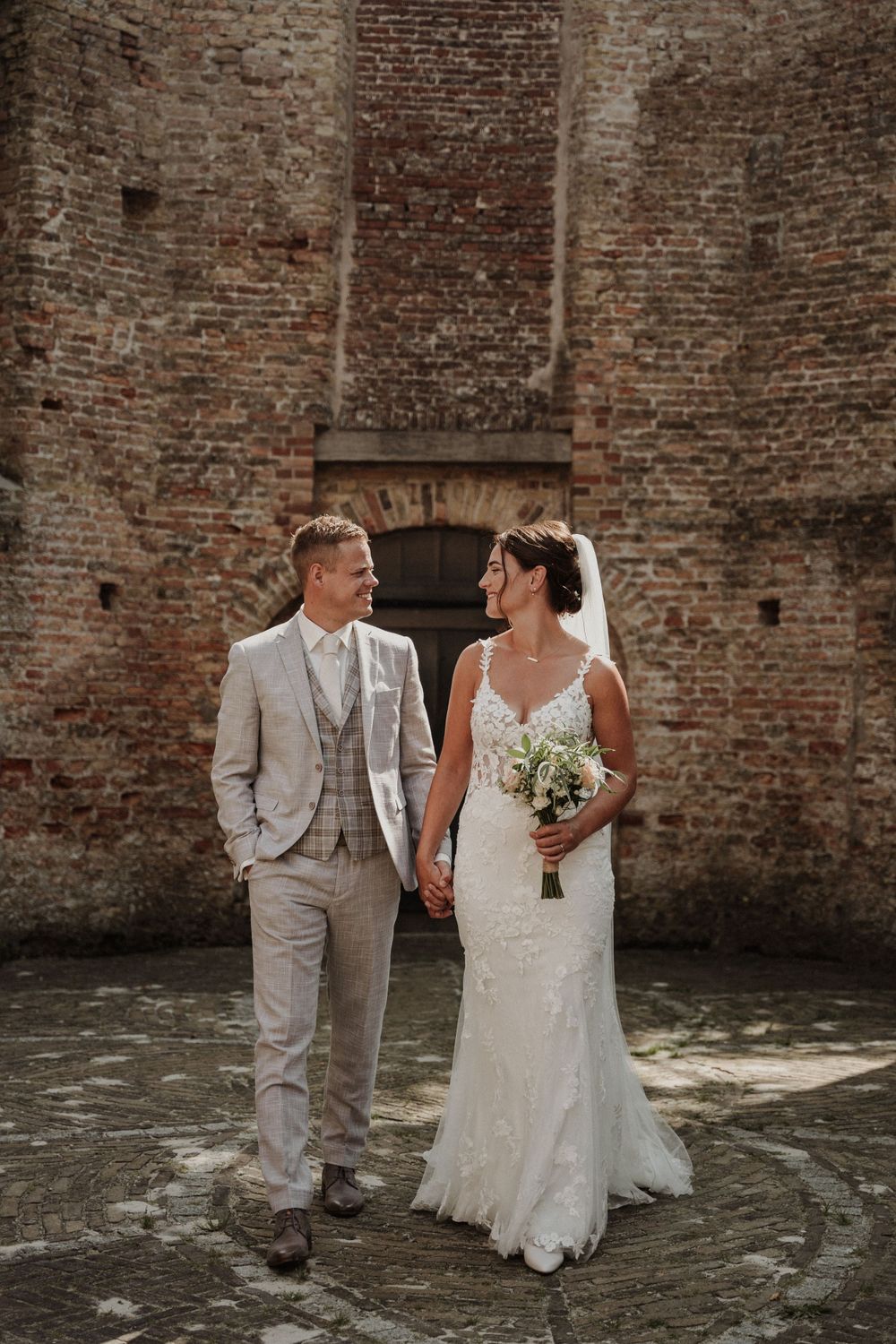 A couple in wedding attire walk hand in hand against a rustic brick wall backdrop on their special day.