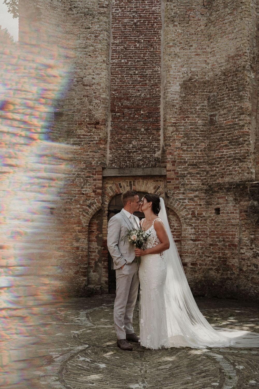 A romantic wedding couple shares a kiss against a rustic brick wall backdrop with a beautiful light prism effect.