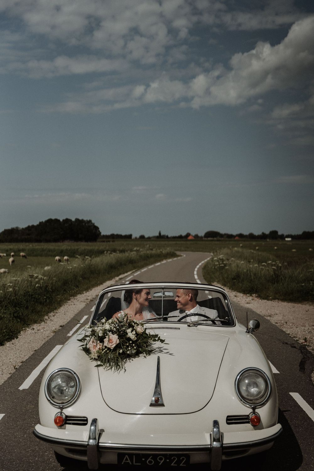 Newlyweds riding in a classic white Porsche convertible on a country road with flowers on the hood.