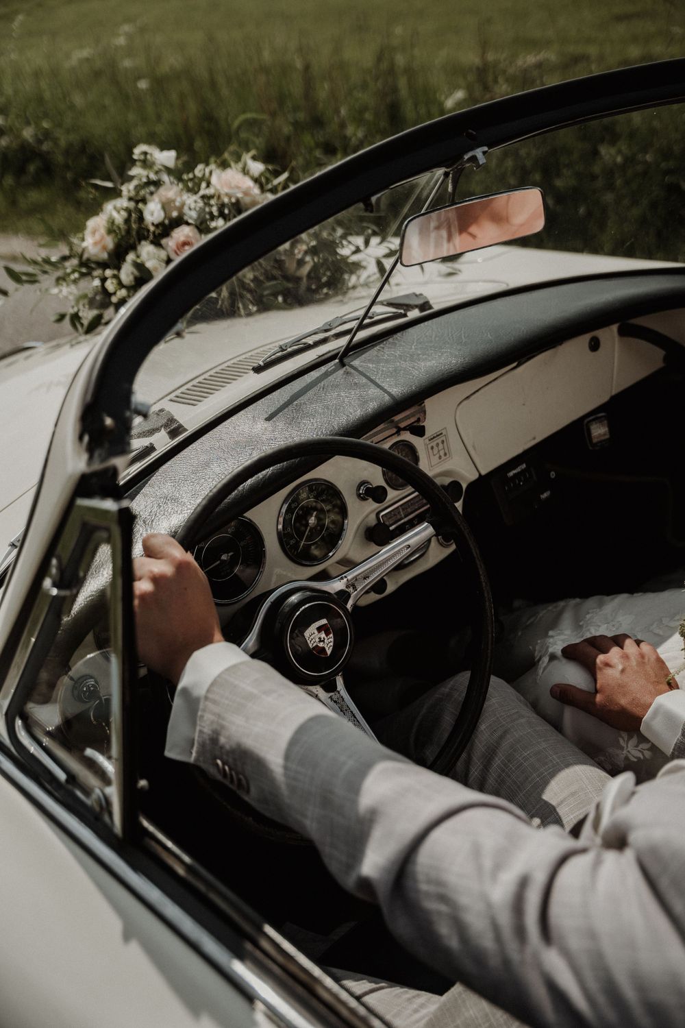 A close-up view of vintage car interior with leather steering wheel and dashboard, showing hands in formal grey attire.