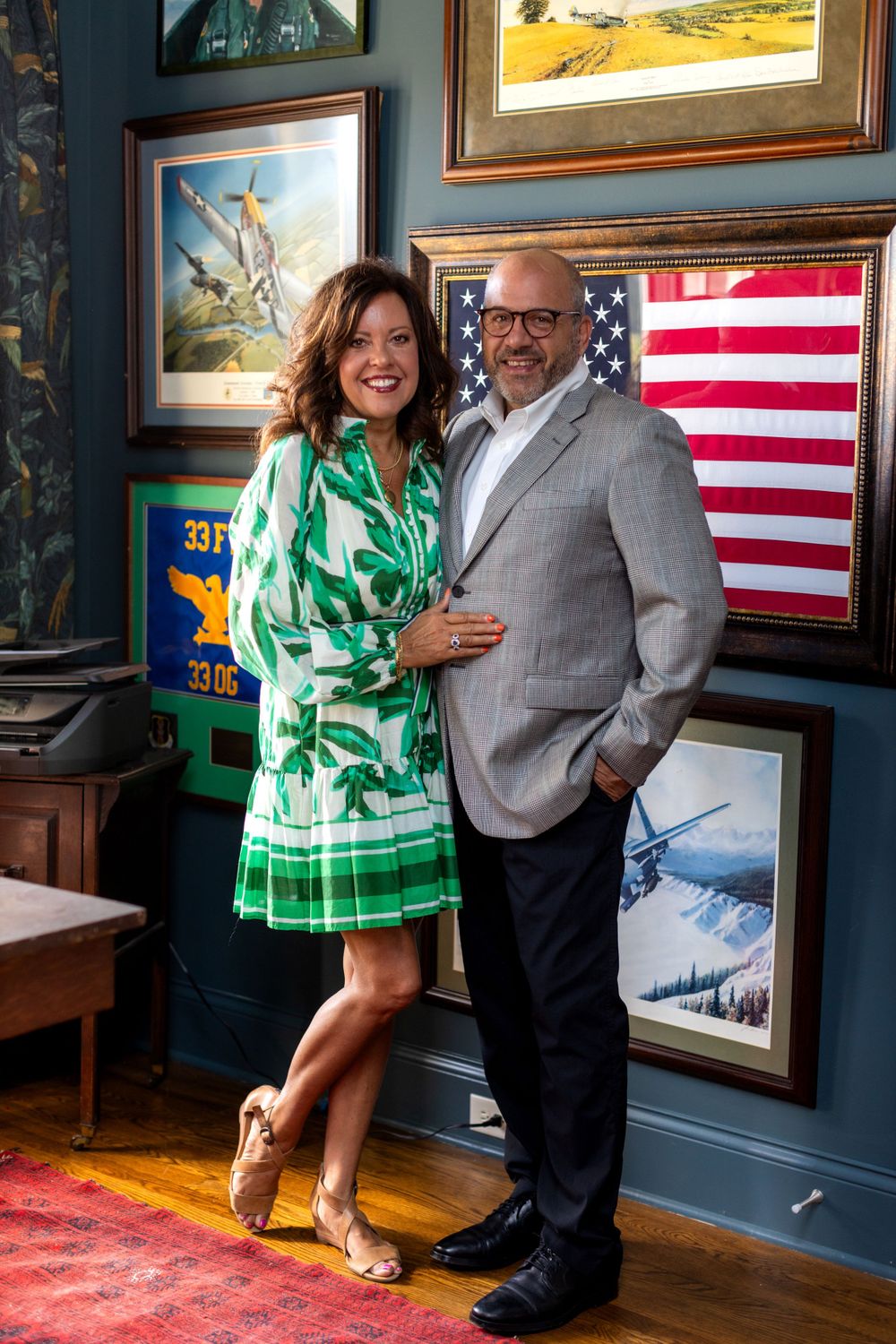 Couple poses together in front of wall art and American flag, wearing green dress and grey suit in elegant interior.