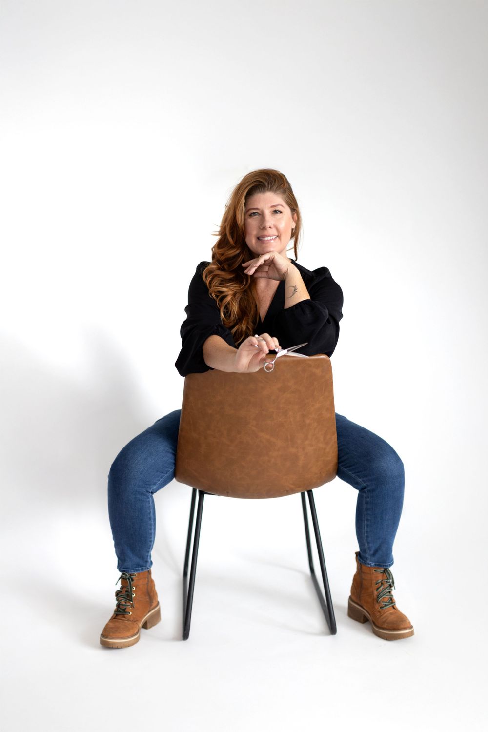 Someone sits casually on a brown chair wearing jeans and boots against a white background.