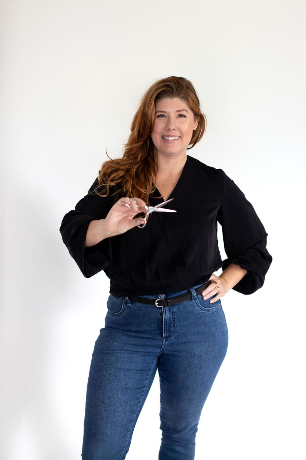 Portrait of someone in a black top and jeans holding scissors against a white backdrop.