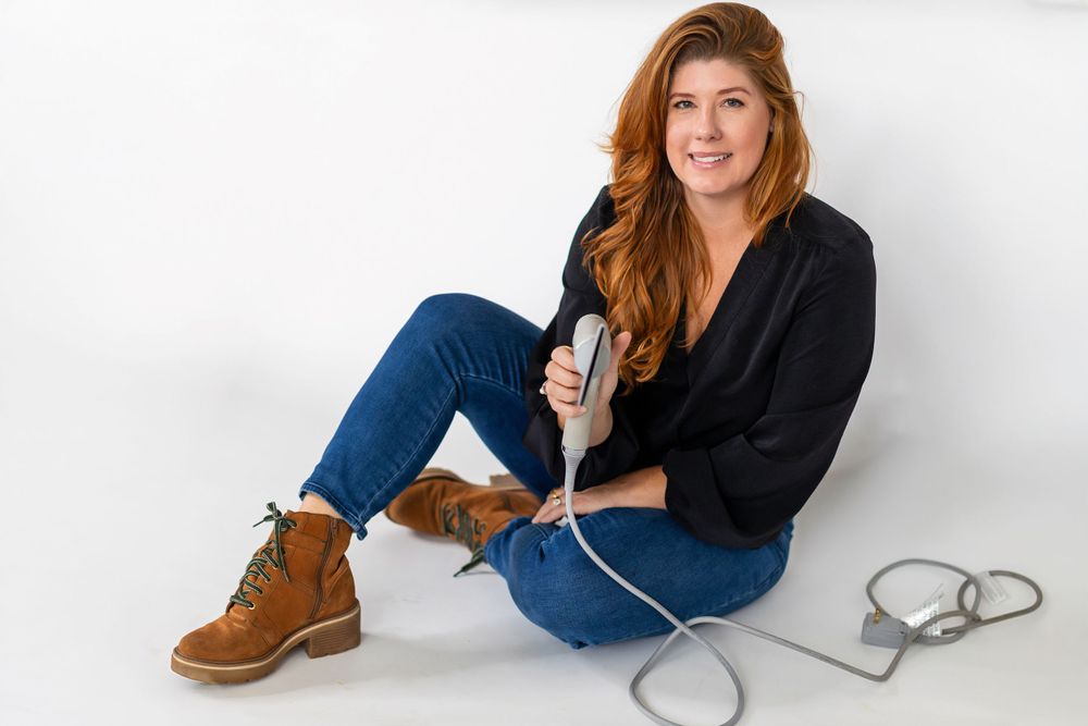 A figure in blue jeans and brown boots sits casually on a white backdrop with a microphone cable nearby.