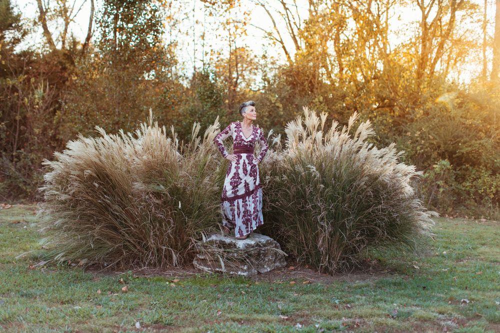 Person in burgundy dress stands on rock between ornamental grasses at sunset.