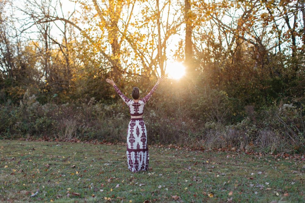 Figure in patterned dress raises arms triumphantly against sunset in outdoor setting.