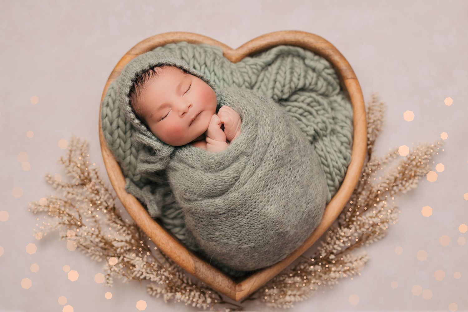 A sleeping newborn swaddled in gray wrap lies in heart-shaped wooden bowl with dried flowers.