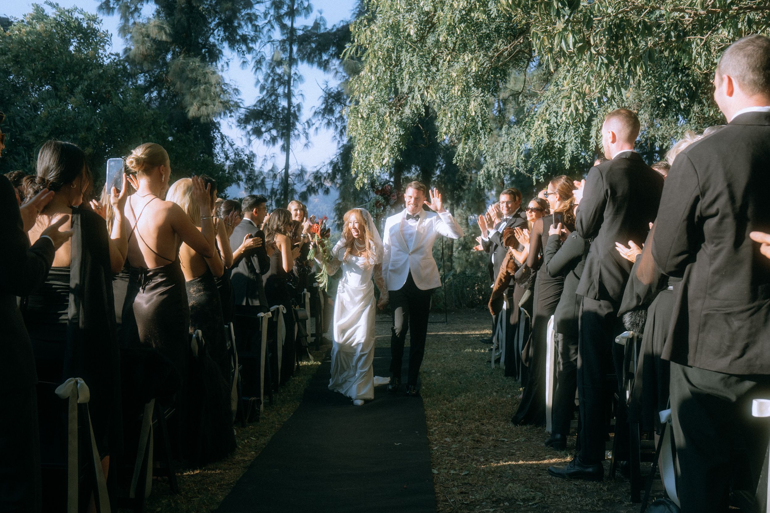 Wedding couple walks down a flower-lined aisle between rows of guests during an outdoor ceremony.