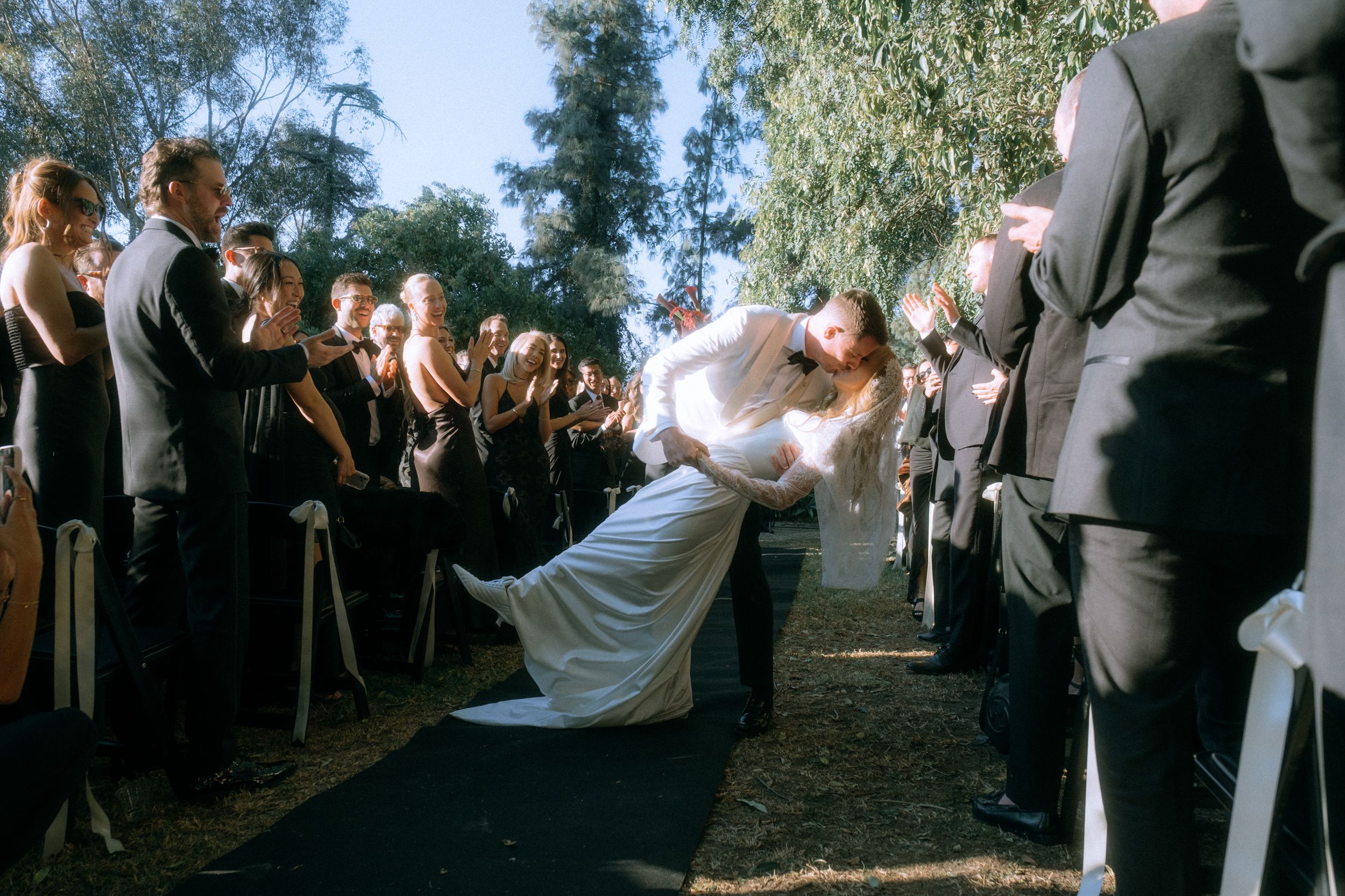 Romantic dip kiss between newlyweds as guests look on during outdoor wedding ceremony.