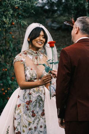 Bride in floral wedding dress holds red rose while sharing joyful moment with partner.