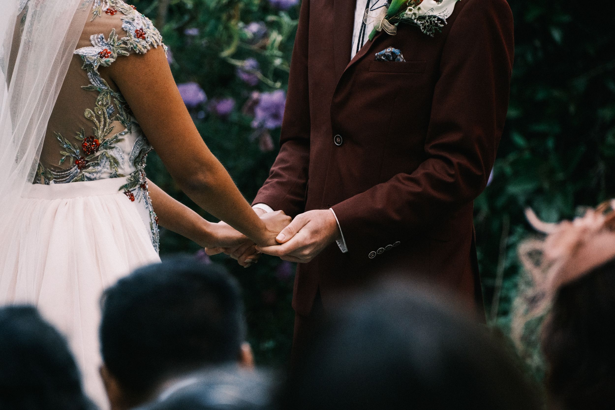 Close-up of couple holding hands during wedding ceremony in floral dress and brown suit.