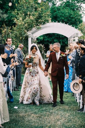Wedding couple exits under white garden arch while guests celebrate with flower petals.