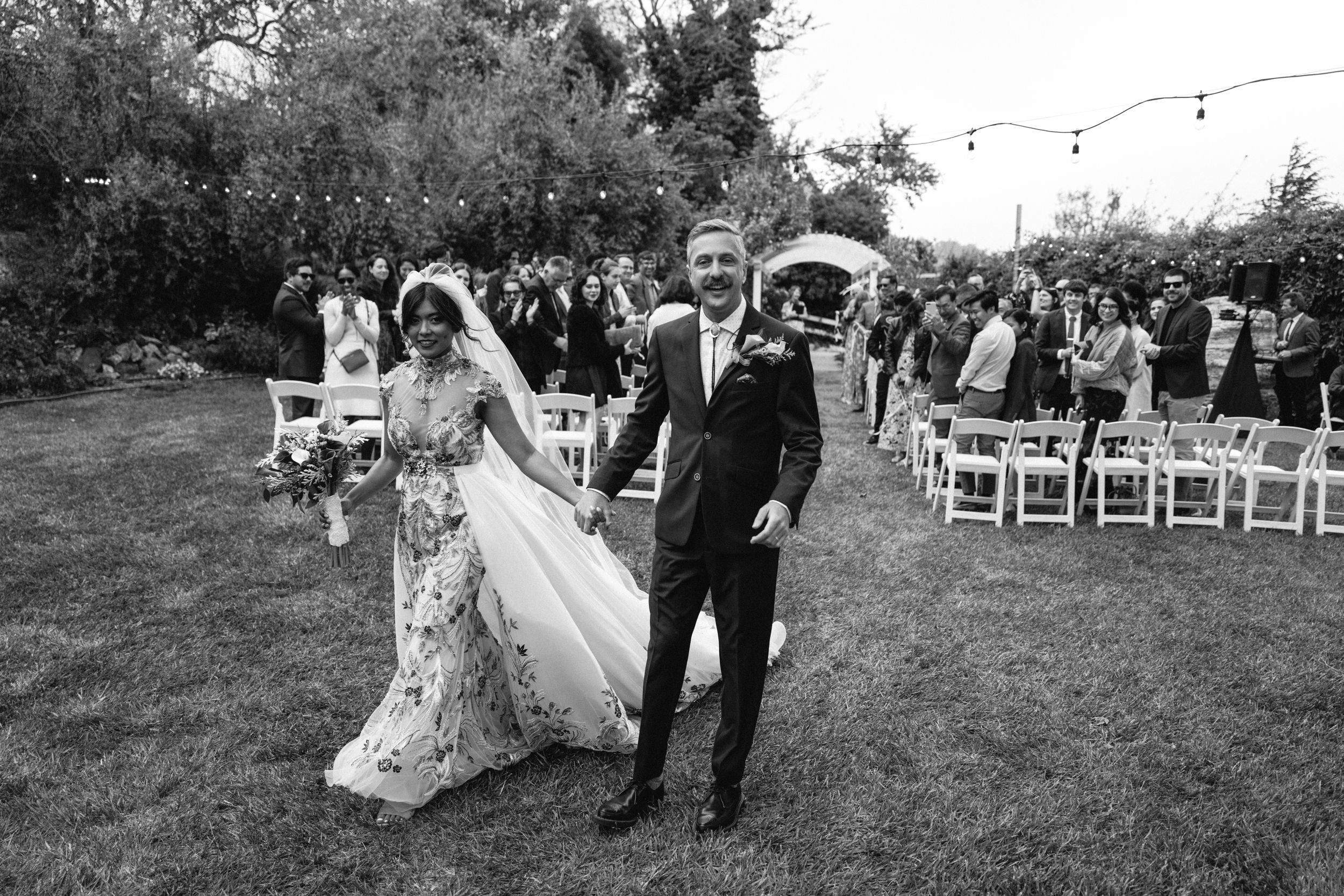 Newlyweds walk hand in hand through garden setting in black and white photography.