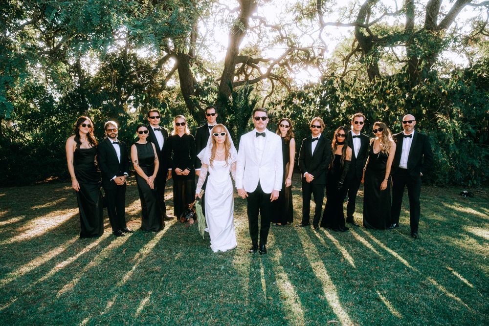 Wedding party in black and white attire poses under sunlit trees.