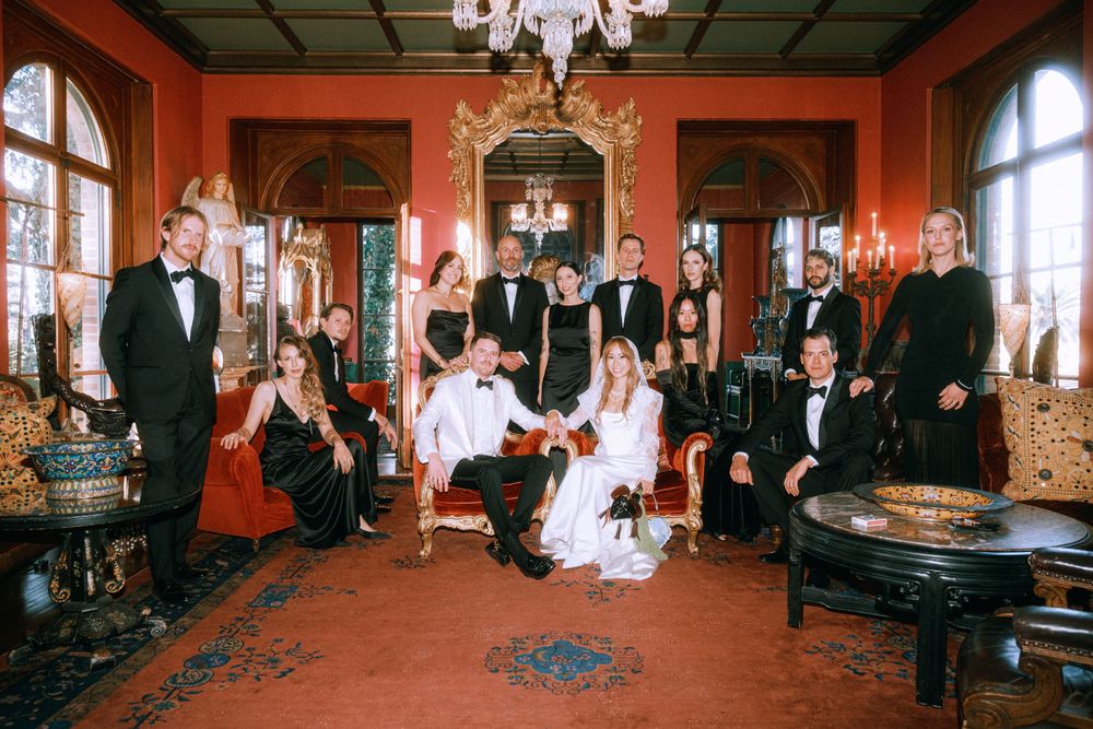 Wedding party gathered in ornate red room with vintage furnishings and chandelier.