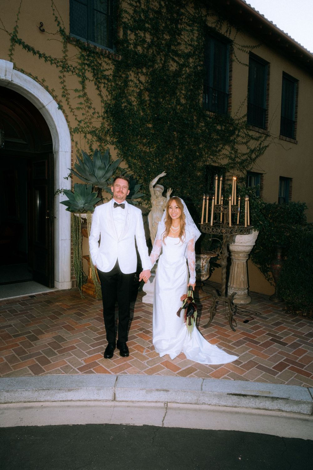 Newlyweds pose on stone steps in front of ivy-covered building.