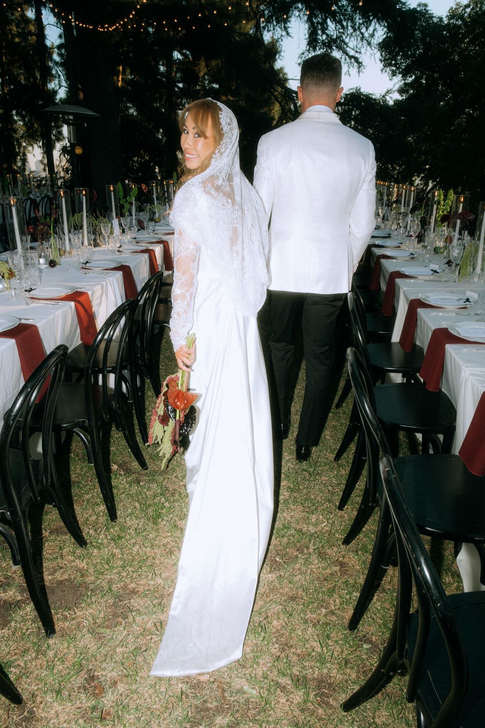 Wedding reception setup with long tables, black chairs and string lights in outdoor garden setting.