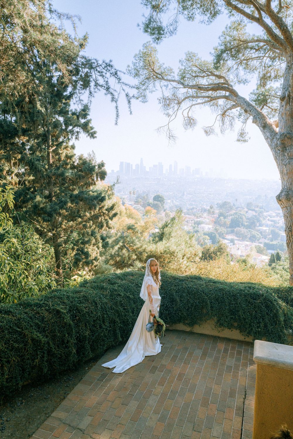 Bride stands on terrace overlooking city skyline through trees at golden hour.